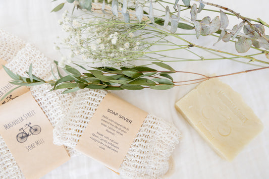 Bar of natural soap with seed paper label, soap saver, and eucalyptus branches on a white cloth.