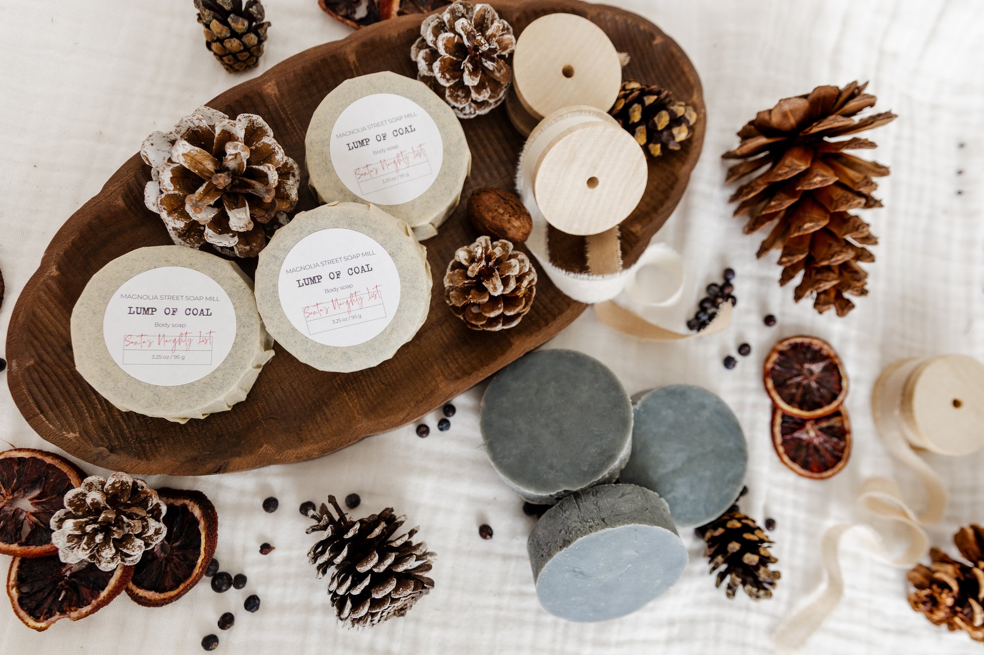 stacks of labeled and unlabeled "Lump of Coal" soap pucks decorated with pine cones, ribbons juniper berries and dried orange slices.