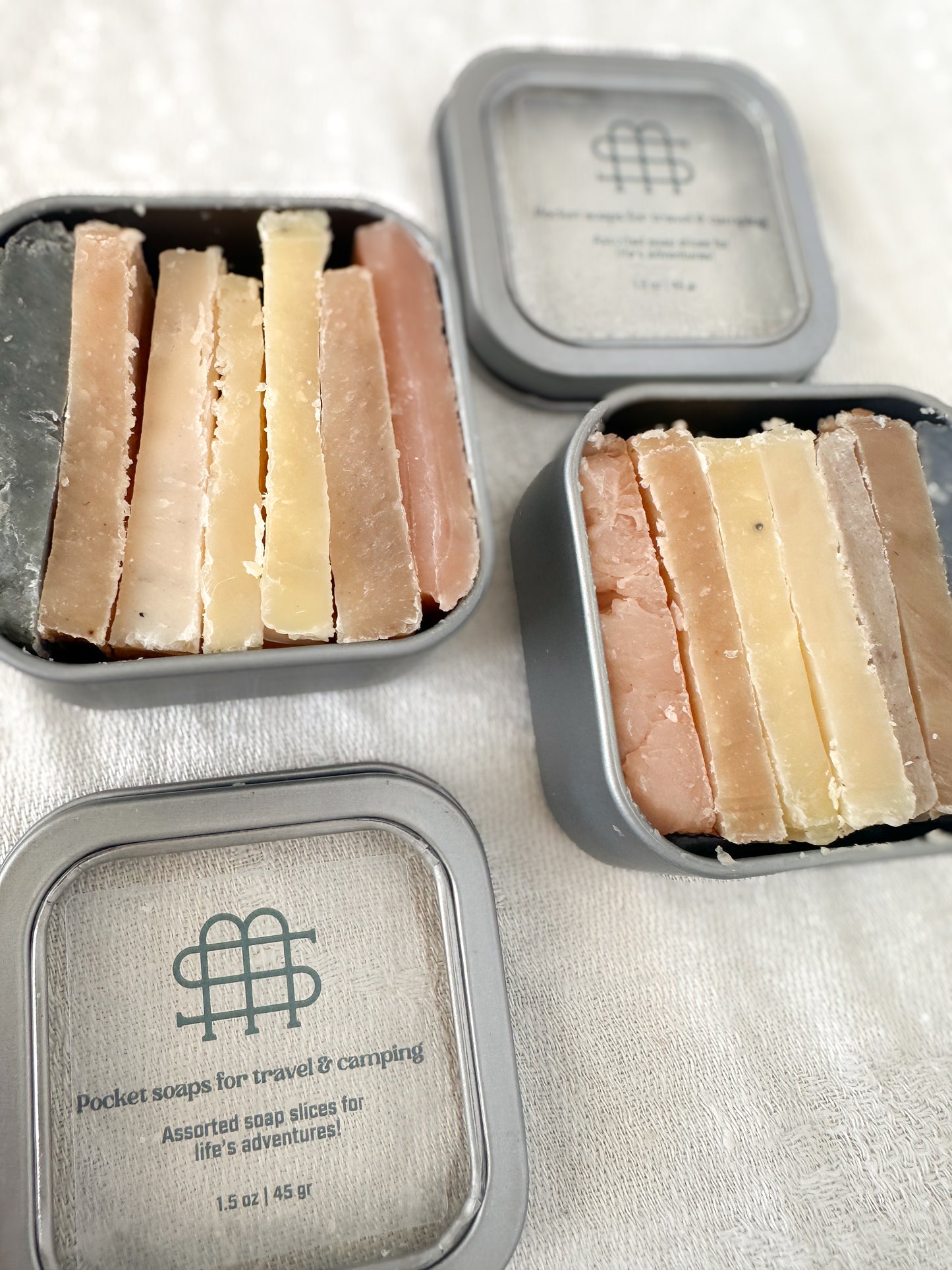 Three containers of assorted soap slices with a brand logo on a light background