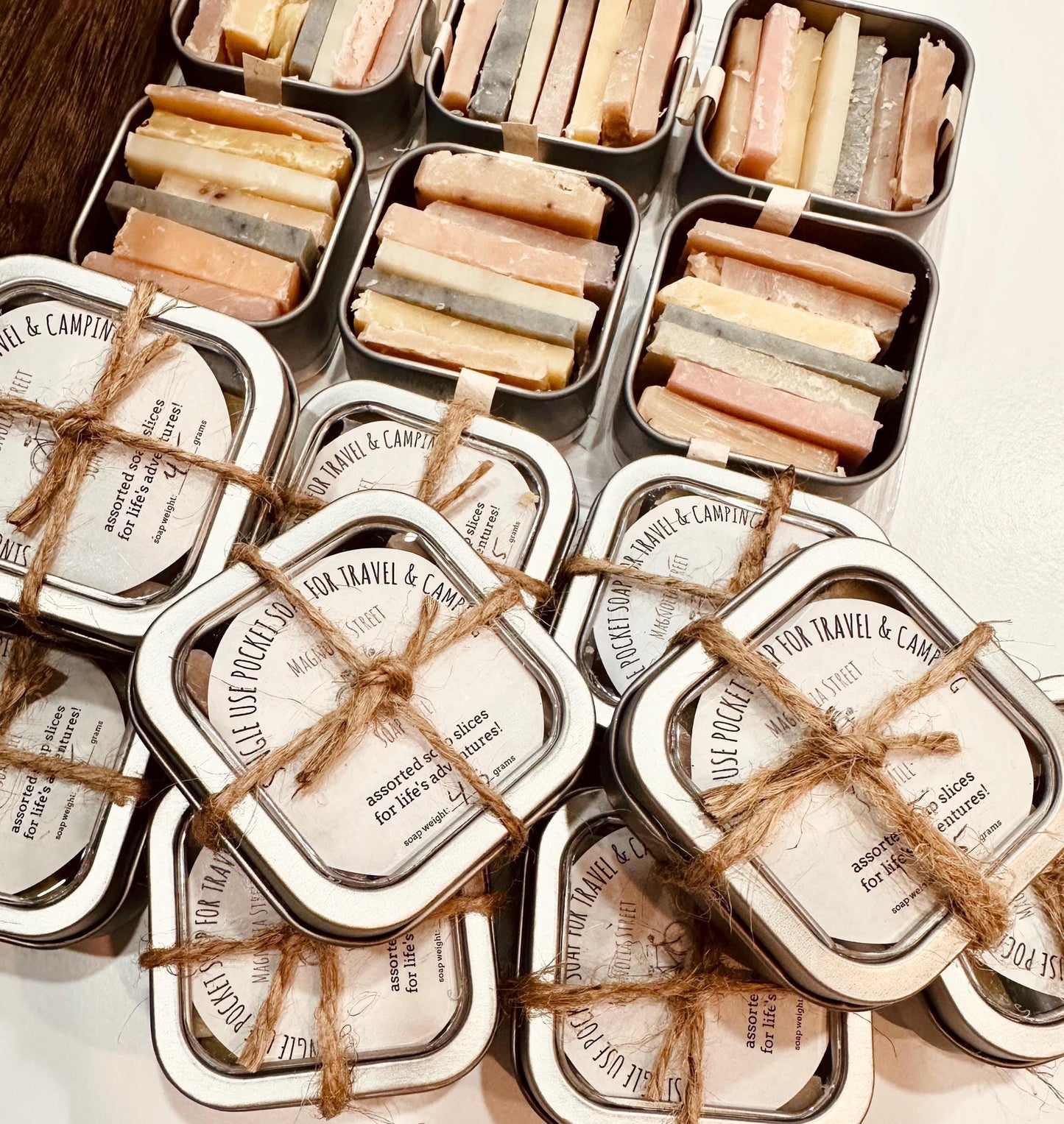 A collection of reusable tins containing pocket soaps with various natural colorants and scents, tied with twine and labeled for travel convenience.