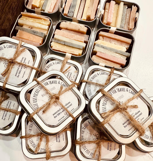 A collection of reusable tins containing pocket soaps with various natural colorants and scents, tied with twine and labeled for travel convenience.