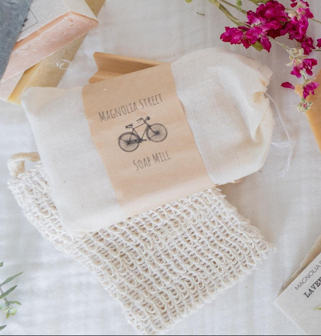 A beige sisal soap saver bag with a drawstring loop, featuring a label that includes the text 'Magnolia Street Soap Mill' and an illustration of a bicycle.