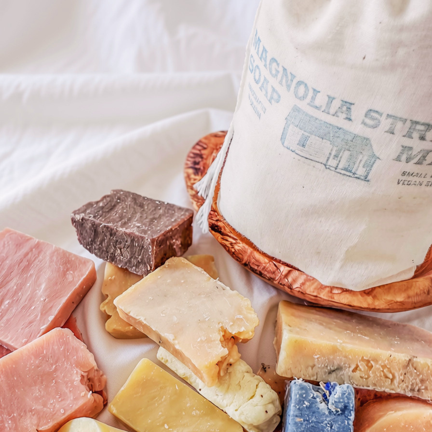 A styled product shot of a white cotton bag labeled 'MAGNOLIA STRIPES SOAP' with a copper-colored base, surrounded by assorted cheese wedges and slices on a white fabric surface.